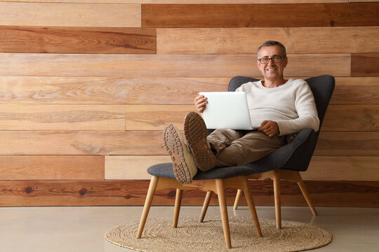Old man sitting in armchair with legs on footstool and using laptop near wooden wall