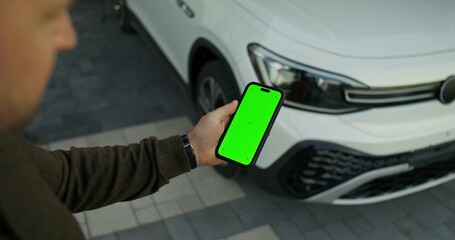 A man holds a mobile phone with green screen in his hand, standing near an electric car. He opens the car with his phone. Close-up of his hands.