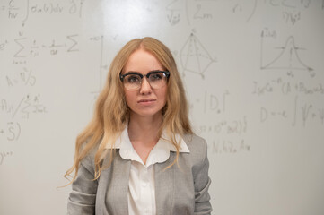 A Caucasian woman stands at a blackboard with written formulas.