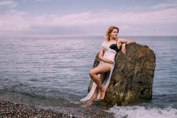A young romantic girl in a white transparent dress stands with her back against a rock in the sea on a cloudy day. She has a smartwatch on her wrist. The concept of a modern goddess