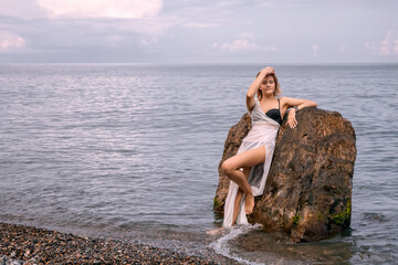 A young romantic girl in a white transparent dress stands with her back against a rock in the sea on a cloudy day. She has a smartwatch on her wrist. The concept of a modern goddess