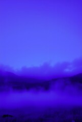 Fototapeta premium A vivid blue hue dominates the landscape, with misty clouds hovering over distant hills. The gradients of color create a serene and ethereal atmosphere.