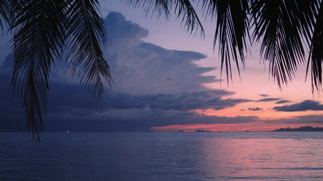 Evening calm sea after beautiful tropical sunset. The remains of beautiful sunset are reflected in the sea water. Silhouettes of palm leaves against the sky. Large thundercloud is approaching the sky