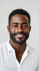 A close-up portrait of a man with a trimmed beard and a warm smile, wearing a button-down shirt and standing against a white background.