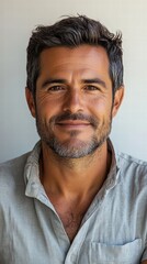 Obraz premium A close-up portrait of a man with a trimmed beard and a warm smile, wearing a button-down shirt and standing against a white background.