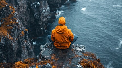 Person wearing orange jacket sitting on cliff edge overlooking the ocean during daytime