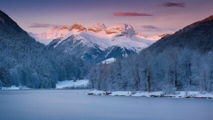 Fototapeta premium Alpine lake reflecting snow-capped mountains at dawn with soft pink and purple hues in the sky surrounded by frosted trees in a winter landscape