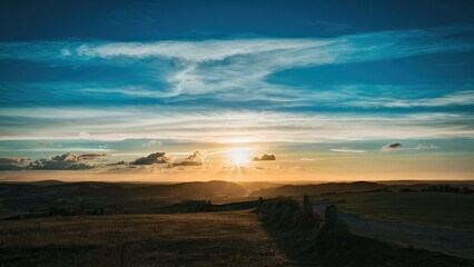 Serene sunset over rolling hills with vibrant orange glow under a deep blue sky featuring wispy clouds and dramatic sunlight streaming through.