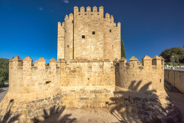 Naklejka premium The Calahorra Tower on a one side of the Roman Bridge in Cordoba town in Spain. Ancient stone tower and walls under a bright blue sky. A historical landmark, possibly a castle or fortress.