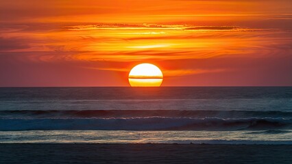 Sunset over tranquil beach with vibrant orange hues reflecting on the calm ocean waves under a clear sky, creating a serene atmosphere