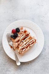 A piece of a puff pastry cake decorated with berries on a white stone background. A piece of a Russian cake decorated with berries on a plate. A piece of the Napoleon cake on a plate top view.
