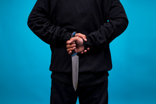Close-up Rear View Of Murderer Holding Knife In Back Over Blue Background 