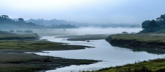 Obraz premium Serene foggy river landscape with calm blue waters reflecting a misty sky surrounded by green wetlands and distant trees in soft focus.
