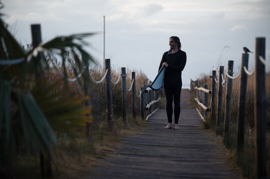 Surfer walking on wooden path at sunset holding surfboard