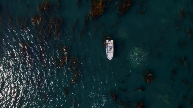 Top down drone ascend of a boat anchored near Cedros Island at midday surrounded by deep blue waters and rocky outcrops or seaweed floating forests