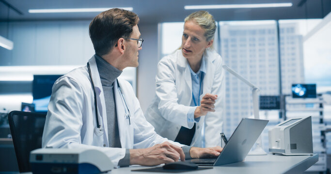 Two Doctors in a.Hospital Office Analyze Patient Data on a Laptop, Discussing Diagnosis and Treatment Options in a Modern Hospital Focused on Advanced Healthcare and Successful Healthcare