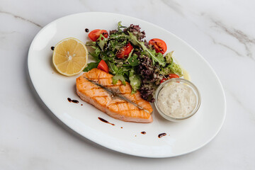 Grilled salmon fillet with fresh salad, lemon slice, tartar sauce, and balsamic glaze drops, placed on a marble table, creating a delicious and healthy meal. Restaurant or cafe menu, cookbook recipe