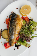 Grilled mackerel fillet is served on a white oval plate with grilled vegetables, fresh salad, lemon slice, and balsamic sauce on a marble table. Restaurant or cafe menu, cookbook recipe