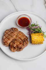 Grilled steak resting on a white plate, accompanied by vibrant corn, red onion, and microgreens, drizzled with barbecue sauce, captured from above. Restaurant or cafe menu, cookbook recipe