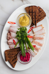 Thinly sliced ukrainian salo served with mustard, horseradish, rye bread, green onions, dill and red chili peppers arranged on white oval plate. Restaurant or cafe menu, cookbook recipe