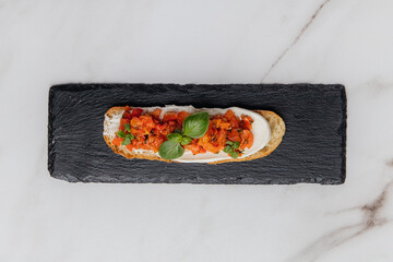 Delicious bruschetta with cream cheese, chopped tomatoes, basil, and spices served on a black slate plate on a marble table, presenting culinary experience. Restaurant or cafe menu, cookbook recipe