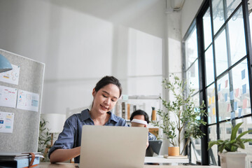 Beautiful young female Asian worker is happy working, using laptop, smiling at desk, thinking of ideas, and concentrating creative marketing jobs in daytime casual office and startup SME business.