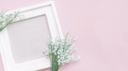 A white wooden picture frame with a fabric-covered center, decorated with delicate lily-of-the-valley flowers on a soft pink background. top view. copy space. banner
