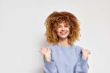 Smiling young woman with curly hair in a light blue sweater on a white background, expressing joy and positivity with hands raised, capturing a cheerful vibe of happiness