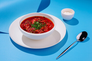 A bowl of red Ukrainian borscht is garnished with fresh herbs, placed on a white plate. A small dish of cream and a silver spoon rest nearby on a bright blue surface.