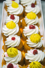 A tray of caramel-colored desserts topped with whipped cream, raspberries, and yellow meringue. The vibrant colors and intricate toppings make it visually appealing.