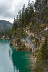 A scenic mountain hiking trail along a rocky cliff overlooking a turquoise alpine lake. The path is lined with wooden railings, surrounded by lush evergreen trees.