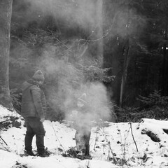 Obraz premium A man and child stand near a small fire in a winter woodland. Smoke obscures them slightly, creating a cold, atmospheric scene. The snow covers the ground.