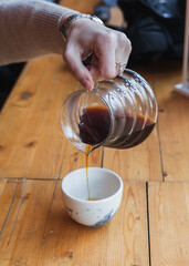 Barista pouring filter coffee from kettle to a ceramic white cup. female or man hand holding glass teapot server with black pour over coffee. alternative brewing, specialty v60. background light wood