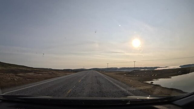 Driving through the tundra in northern Norway on the E69 road. The landscape features plains and hills covered with moss and lichen, with patches of snow. The sun shines directly ahead.