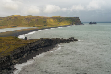 Vue panoramique sur la plage de sable noir de Reynisfjara en Islande, avec ses falaises abruptes et les formations rocheuses de Reynisdrangar émergeant de l’Atlantique sous une lumière dramatique.