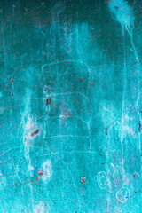 A close-up of a weathered turquoise metal surface with scratches, rust spots, and faded markings. The textured appearance shows signs of wear and oxidation, creating an abstract pattern