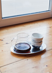 Glass server with black filter coffee inside and ceramic cup stand on wooden cafeteria table, day light from window. Making filtered coffee in coffee shop. Pour over method of alternative brewing, v60