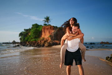 Joyful couple enjoys piggyback ride on tropical beach at sunset. Man carries girlfriend, both...