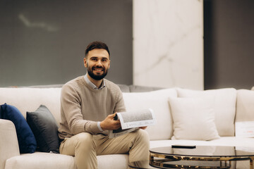 Obraz premium Confident male expert presenting variety samples of upholstery fabric in dark lightened showroom. Portrait of smiling brunet salesman showing colorful textile palette. Concept of interior design.