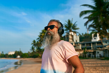 Grey-bearded senior man enjoying music on tropical beach. Stylish elder in sunglasses and wireless...