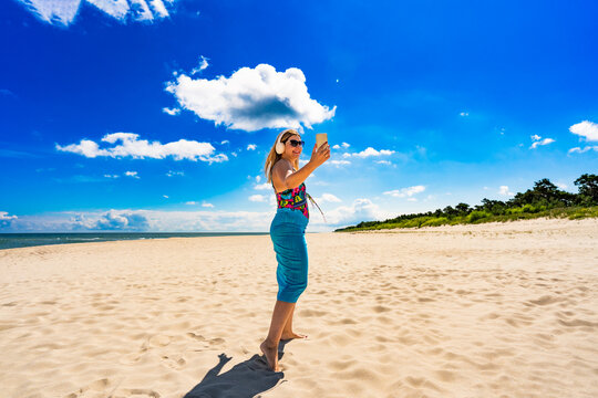 Summer holidays on beach with music. Portrait of beautiful middle aged woman listening to music in headphones holding smartphone on sandy beach. Front view