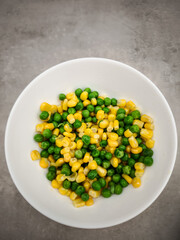Small white ceramic bowl containing steamed green peas and golden corn kernels, resting on neutral grey surface copy space Galentines day
