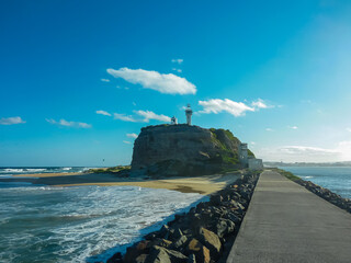 Nobbys Lighthouse perched atop rugged cliffs at Newcastle Beach, New South Wales, Australia. Concrete breakwater path leads to historic lighthouse. Pacific Ocean waves crash against rocky shoreline