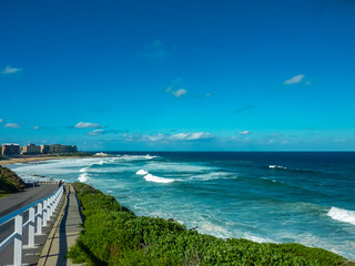 Scenic coastal walkway along Newcastle Beach, NSW, Australia, featuring turquoise ocean waves, white railings, and lush green vegetation. Modern beachfront buildings line the horizon