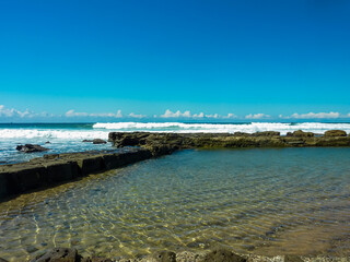 Crystal-clear tidal ocean pool bordered by dark volcanic rocks at Bar Beach, Newcastle, Australia. Turquoise waves crash against rocky shore under blue sky with white clouds. Serene coastal landscape