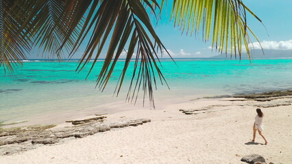 Young woman is enjoying a sunny day, walking on a white sand beach bordered by a turquoise lagoon and lush vegetation