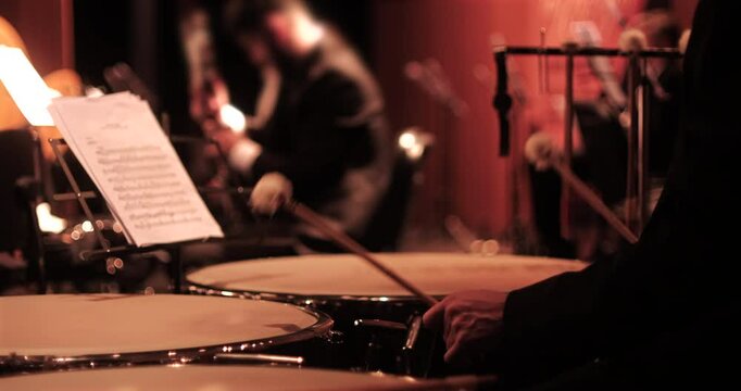 A close-up view of a timpani player with drumsticks poised over the instrument, and sheet music visible on a stand. The blurred background captures other orchestra musicians