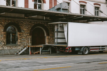 Small transport truck with empty and open container door and lift next to an old building with a loading bay door, no people