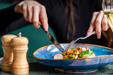 A close-up of a person enjoying a vibrant salad served in a stylish ceramic bowl. The hands are elegantly using a knife and fork, with seasoning shakers in the background.