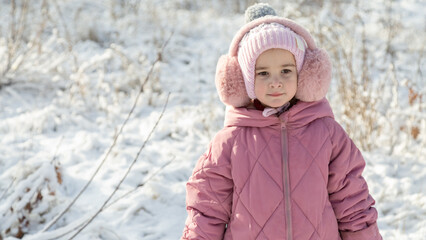 Cheerful positive child girl wearing a outerwear smiling walking through a snowy countryside forest
